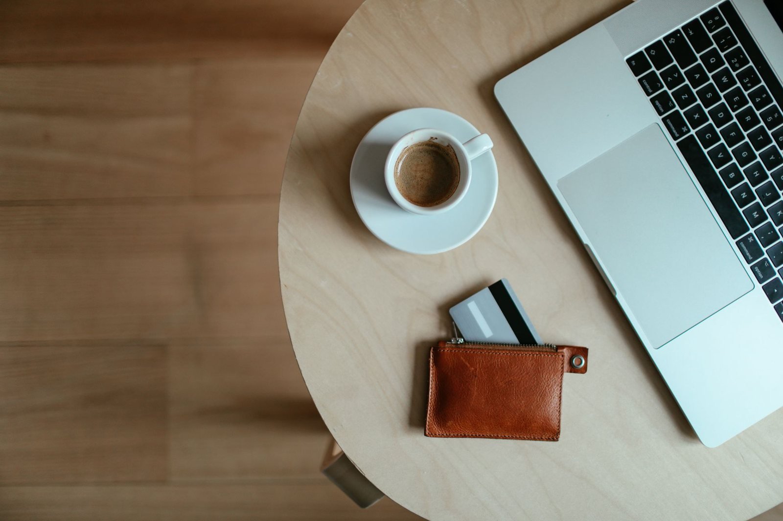A laptop, an espresso, and a leather cardholder with cards on a round wood table