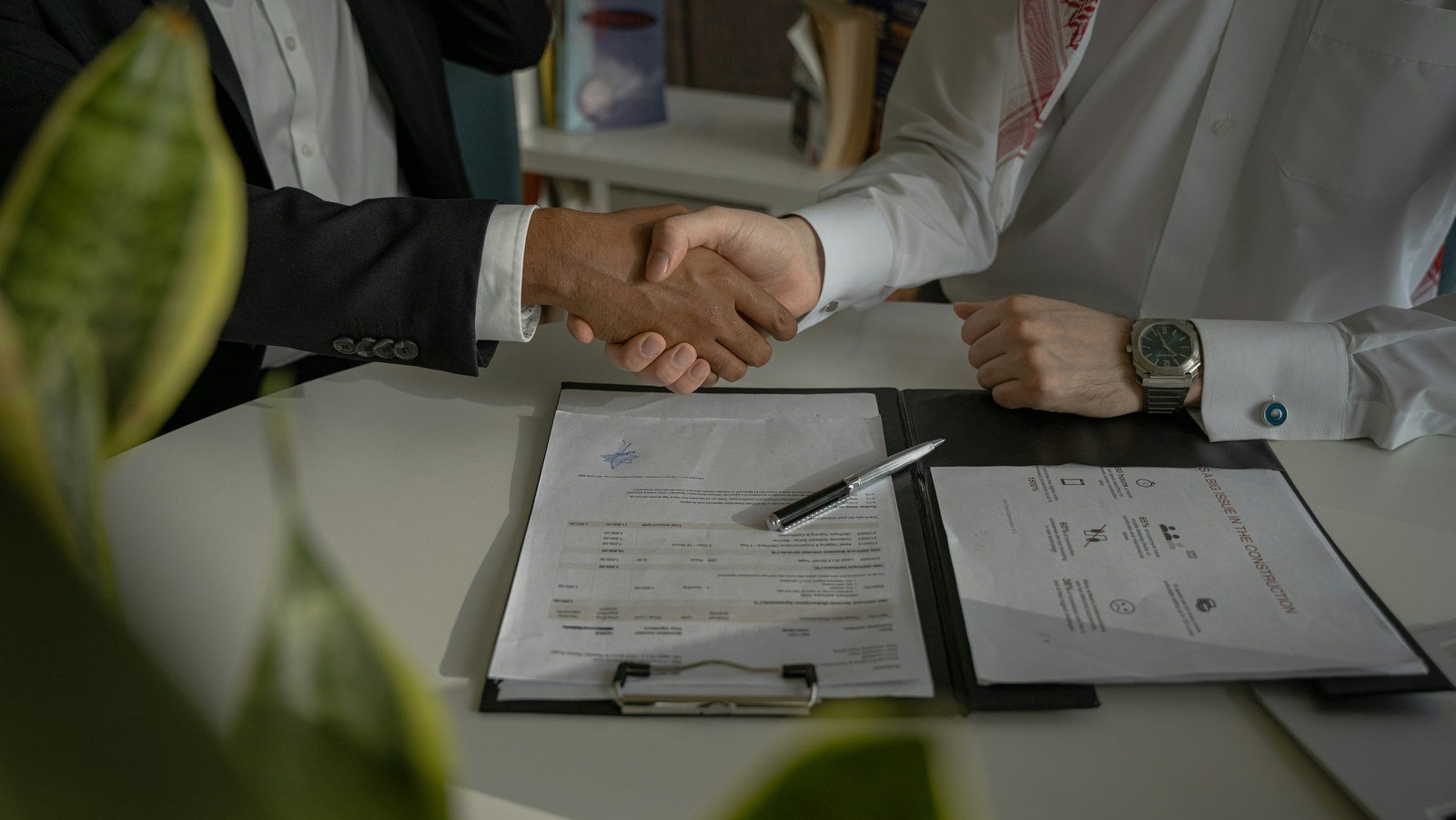 Two people shaking hands over paperwork and a pen on a desk